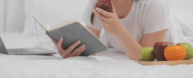 Efficiente giovane ragazza millenaria seduta su un letto al mattino usa il computer portatile e mangia croissant e beve caffè a colazione