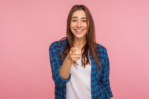 Ehi, abbiamo bisogno di te. Ritratto di ragazza carina allegra in camicia a scacchi con un sorriso a trentadue denti che punta alla telecamera, indicando il dito e felice di fare una scelta. studio indoor girato isolato su sfondo rosa