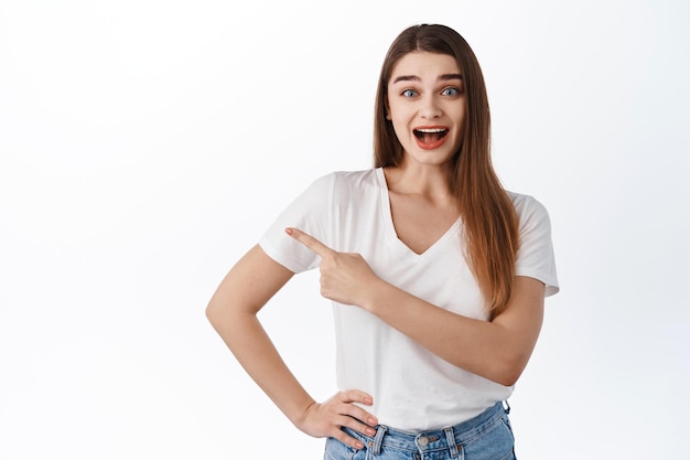 Ehi, dai un'occhiata. Eccitato bella ragazza sorridente stupita, puntando il dito da parte a copyspace, mostrando pubblicità, notizie fantastiche, in piedi in t-shirt su sfondo bianco