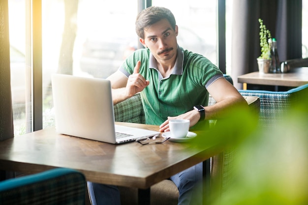 Ehi, giovane uomo d'affari serio in maglietta verde seduto e lavorando sul laptop guardando la fotocamera con segnale di avvertimento business e concetto di freelance tiro al coperto vicino alla grande finestra durante il giorno