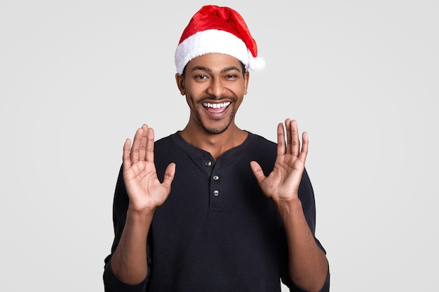 Ehi, ragazzi, Babbo Natale viene da voi! Felice maschio dalla pelle scura positivo con ampio sorriso, solleva le mani e mostra i palmi, indossa un cappello festivo, modelli su bianco. Vacanze ed emozioni.