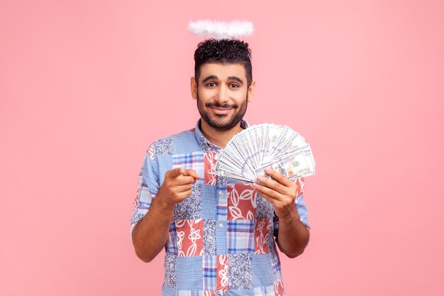 Ehi tu, fai soldi. Ritratto di uomo barbuto angelico positivo con alone sopra la testa in camicia blu che tiene un ventaglio di banconote da un dollaro, puntando il dito verso la fotocamera. Studio indoor girato isolato su sfondo rosa.