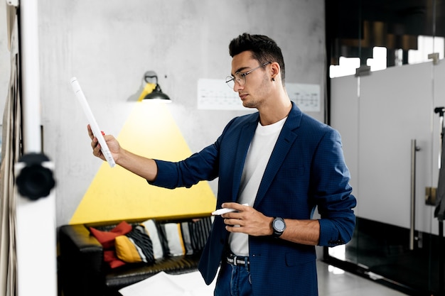 Elegante architetto vestito con giacca a scacchi blu e jeans lavora con progetti accanto a una tavola sul muro nell'ufficio moderno.