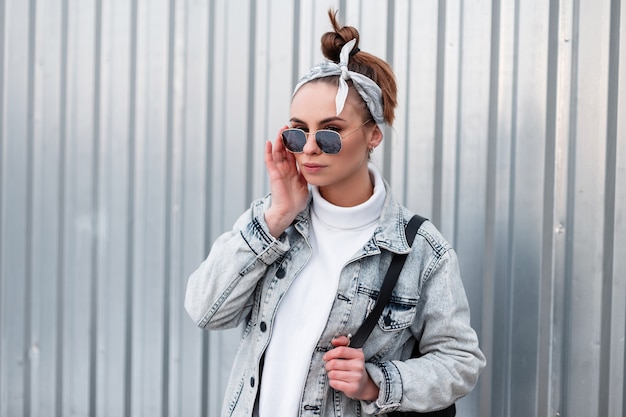 Elegante attraente giovane donna hipster in una bandana alla moda in occhiali da sole alla moda in una giacca estiva con uno zaino in un maglione vicino a una parete di metallo.