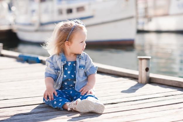 Elegante bambina che indossa abiti in denim alla moda seduta sul molo di mare in legno all'aperto