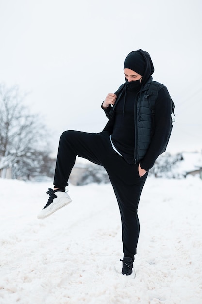 Elegante bel giovane in tuta sportiva nera alla moda con una maschera protettiva e uno zaino cammina con le scarpe da ginnastica nella neve in una giornata invernale