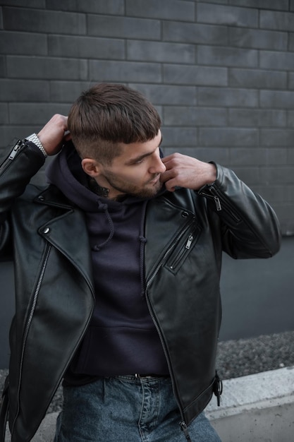 Elegante bel modello hipster ragazzo con i capelli in un vestito alla moda con una giacca di pelle e felpa con cappuccio indossa un cappuccio per strada vicino a un muro di mattoni neri