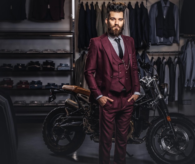 Elegante bell'uomo vestito con abito rosso vintage in posa con le mani in tasca vicino a una moto sportiva retrò al negozio di abbigliamento maschile.