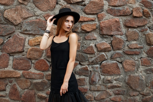 Elegante bella donna in abiti neri alla moda con il cappello vicino a un muro di pietra