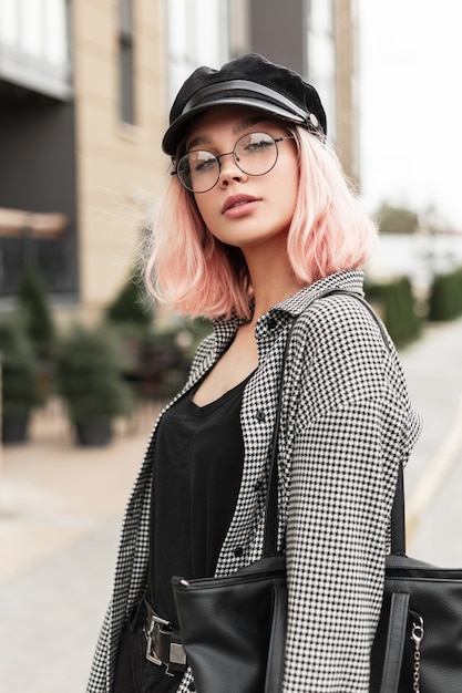 Elegante bella giovane donna con gli occhiali e un copricapo in una maglietta nera con una camicia di moda e una borsa cammina per strada