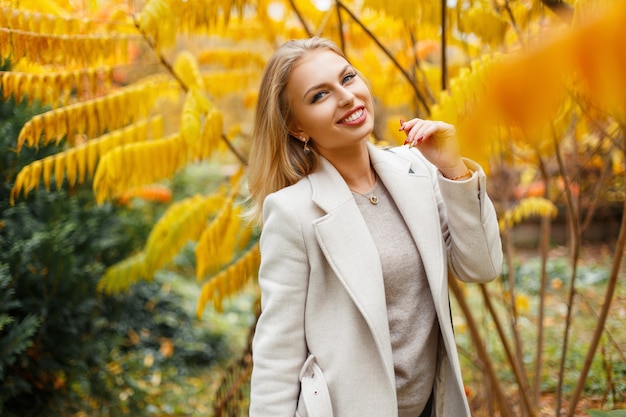 Elegante bella giovane donna con un sorriso in un cappotto autunno alla moda con una borsa vicino alle foglie gialle autunnali