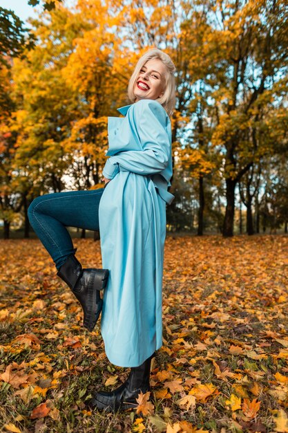 Elegante bella giovane donna felice in un cappotto blu alla moda e scarpe di pelle con jeans sta riposando nella natura con alberi autunnali con fogliame autunnale giallo brillante