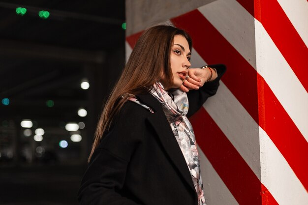 Elegante bella giovane donna in abiti eleganti della gioventù in posa sul parcheggio coperto. Bella modella carina in cappotto nero con una sciarpa di seta vintage si trova vicino a una colonna con una linea rosso-bianca all'aperto.