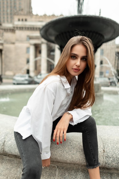 Elegante bella modella di moda donna in abiti casual alla moda con una camicia bianca e jeans neri si siede e posa vicino alla fontana della città