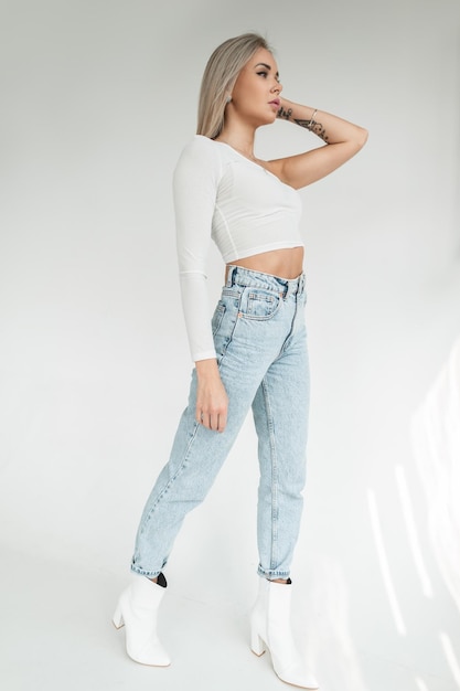 Elegante bella ragazza bionda gentile in una maglietta bianca alla moda e jeans blu vintage a vita alta con scarpe bianche su sfondo bianco in studio