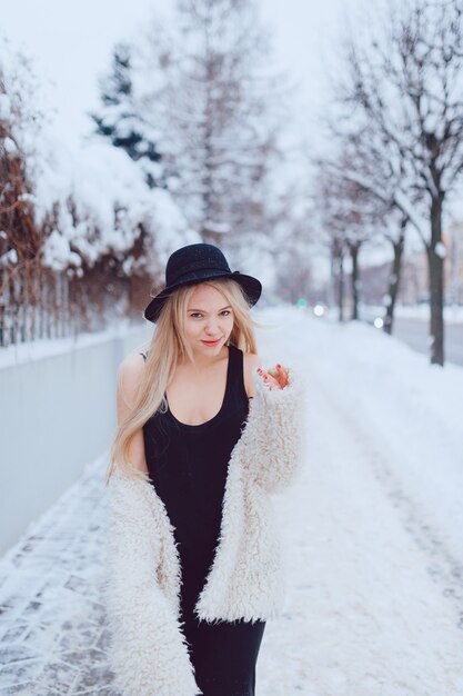 Elegante bella ragazza bionda in cappotto e cappello in posa vicino al recinto in strada