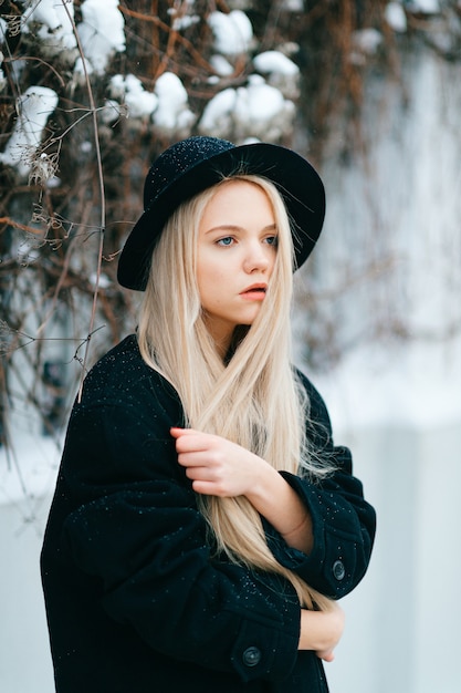 Elegante bella ragazza bionda in vestiti neri e cappello in posa vicino al recinto in strada