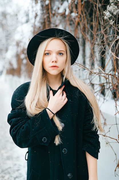 Elegante bella ragazza bionda in vestiti neri e cappello in posa vicino al recinto in strada