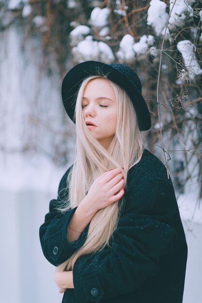 Elegante bella ragazza bionda in vestiti neri e cappello in posa vicino al recinto in strada