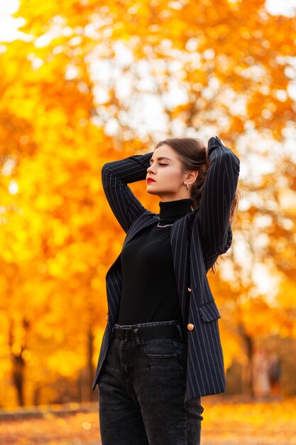 Elegante bella ragazza d'affari di successo in un abito nero alla moda con un maglione vintage e blazer cammina all'aperto su uno sfondo di fogliame autunnale al tramonto