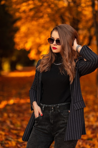 Elegante bella ragazza in abiti alla moda con un blazer e un maglione che indossa occhiali da sole eleganti in un parco autunnale con fogliame arancione