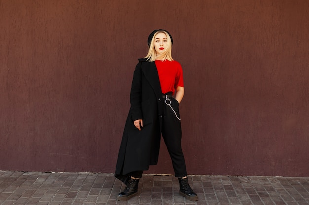 Elegante bella ragazza in un berretto nero e cappotto autunnale con stivali vicino al muro in una giornata autunnale