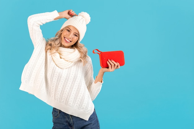 Elegante bionda sorridente bella giovane donna che tiene altoparlante wireless ascoltando musica che indossa un maglione bianco e cappello lavorato a maglia in posa sul blu