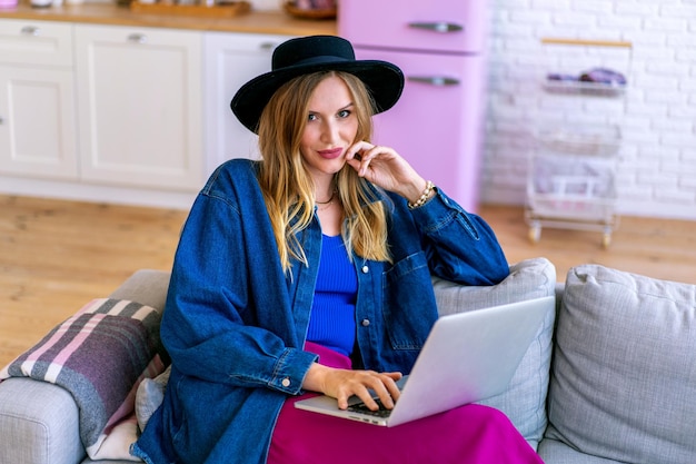 Elegante blogger donna che usa il suo computer portatile, seduta sul divano in soggiorno, ufficio a casa. Abbigliamento di tendenza.