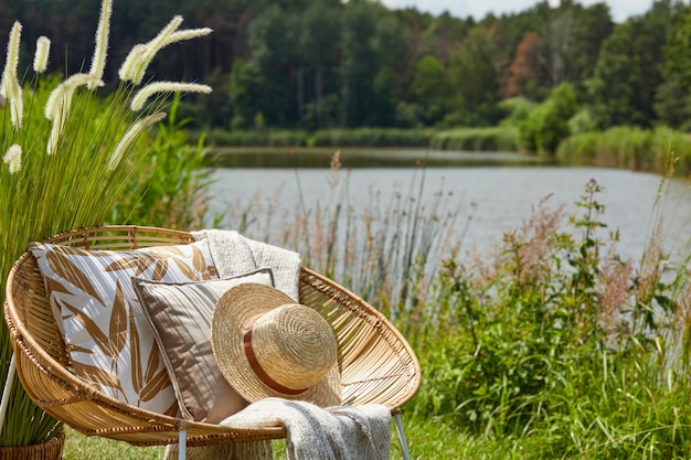 Elegante composizione di giardino esterno sul lago con poltrona di design in rattan, tavolino, cuscini plaid, bevande ed eleganti accessori