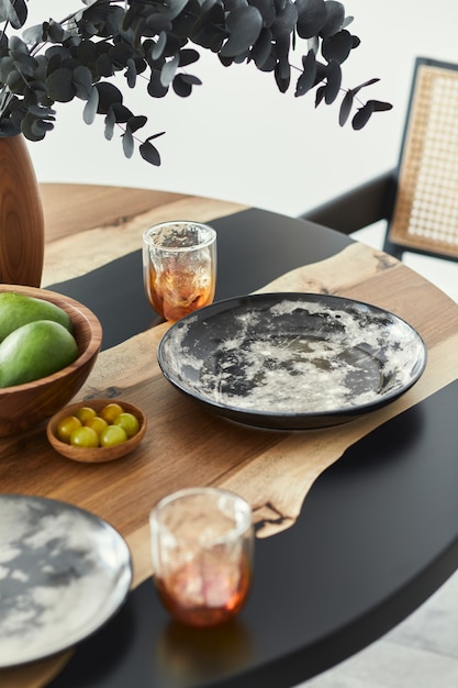 Elegante composizione sul tavolo di design con eleganti piatti, bicchieri, frutta e fiori di eucalipto nero in vaso di legno. Interno moderno della sala da pranzo.