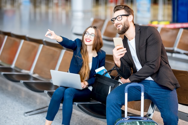 Elegante coppia d'affari seduta con laptop, telefono e valigia nella sala d'attesa dell'aeroporto. Concetto di viaggio d'affari