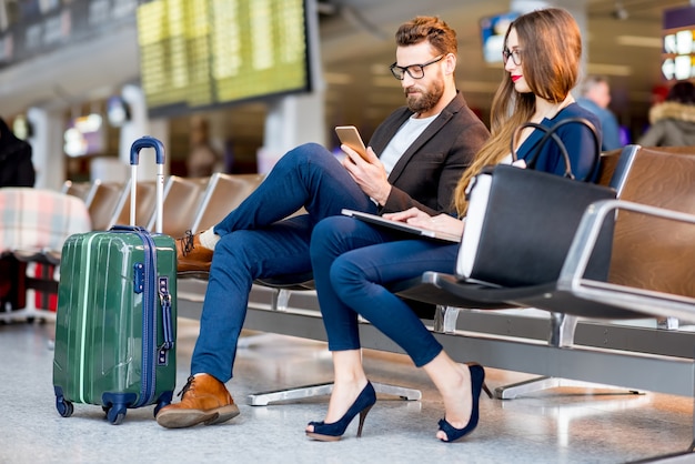 Elegante coppia di affari che si siede con il telefono e prenota nella sala d'attesa dell'aeroporto. Concetto di viaggio d'affari