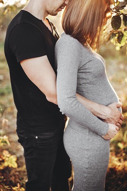 Elegante coppia incinta che si tiene per mano sulla pancia e si abbraccia alla luce del sole nel parco autunnale sotto l'albero Giovani genitori felici mamma e papà che abbracciano il pancione godendosi un bel momento al tramonto