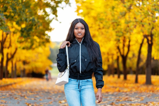 Elegante donna afroamericana felice in giacca nera alla moda e jeans blu con borsa bianca cammina nel parco con incredibili foglie autunnali dorate