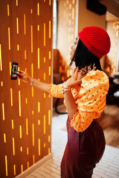 Elegante donna afroamericana in berretto francese rosso, camicetta a pois con grande collo d'oro e pantaloni di pelle conduce una trasmissione in diretta dal cellulare per i social network.