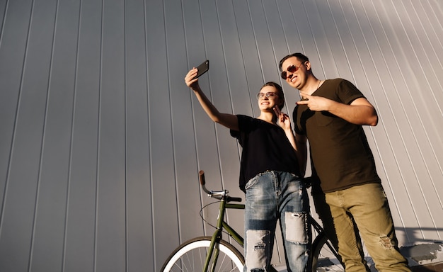 Elegante donna attraente e ragazzo che mostra un segno di pace mentre si fa una foto di selfie, utilizzando un telefono cellulare, in piedi con la bici, all'aperto.