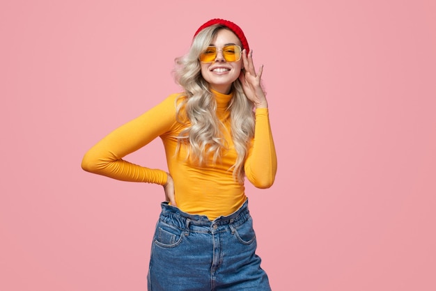 Elegante donna bionda hipster con cappello rosso e dolcevita giallo che tocca occhiali da sole alla moda e guarda la fotocamera su sfondo rosa