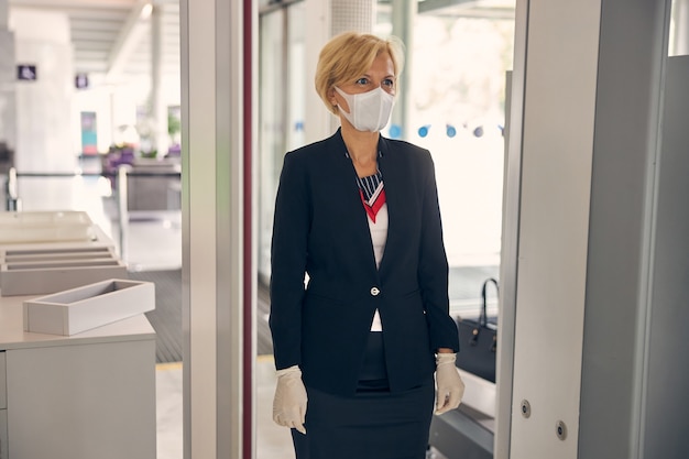 Elegante donna bionda in maschera medica che passa attraverso il cancello di controllo di sicurezza nella sala partenze