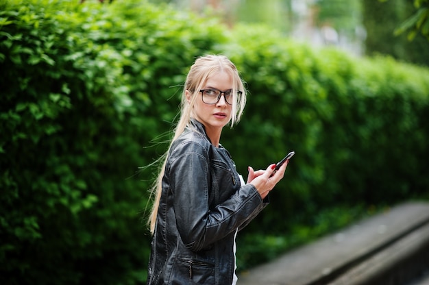 Elegante donna bionda indossare jeans, occhiali e giacca di pelle con il telefono a portata di mano