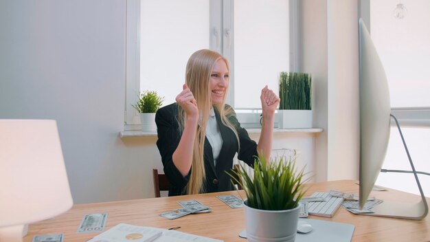 Elegante donna bionda seduta alla scrivania sul posto di lavoro con il computer che tiene in mano un fascio di soldi guarda...