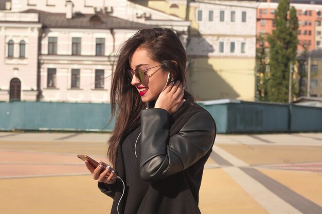 Elegante donna bruna con gli occhiali ascolta musica sul telefono cellulare, in piedi per strada