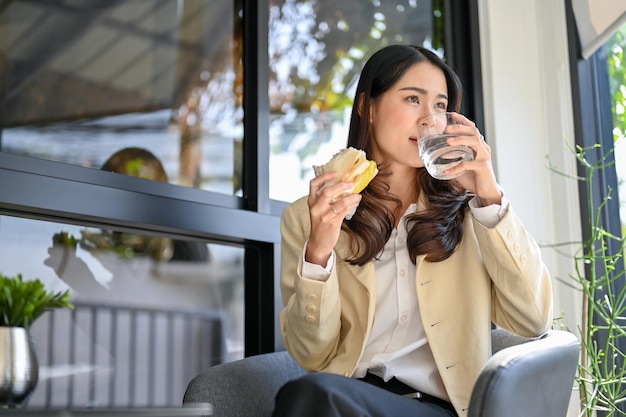 Elegante donna d'affari asiatica che fa colazione al caffè bevendo acqua e mangiando un panino