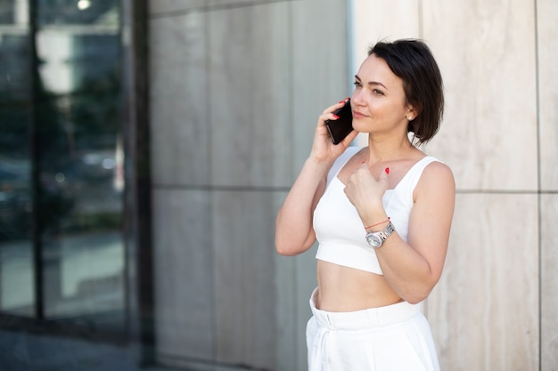 Elegante donna d'affari con orologio da polso che parla al telefono mentre si sta in piedi per strada e guarda lontano