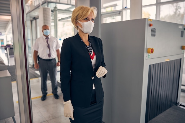 Elegante donna d'affari in maschera medica protettiva che passa attraverso il cancello del check-in mentre un lavoratore aeroportuale maschio in piedi dietro di lei