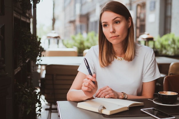 Elegante donna d'affari in un caffè di strada che prende appunti su un taccuino e beve caffè