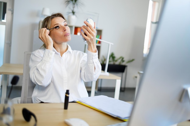 Elegante donna d'affari rinfresca il trucco applicando il mascara mentre è seduto sul posto di lavoro in ufficio