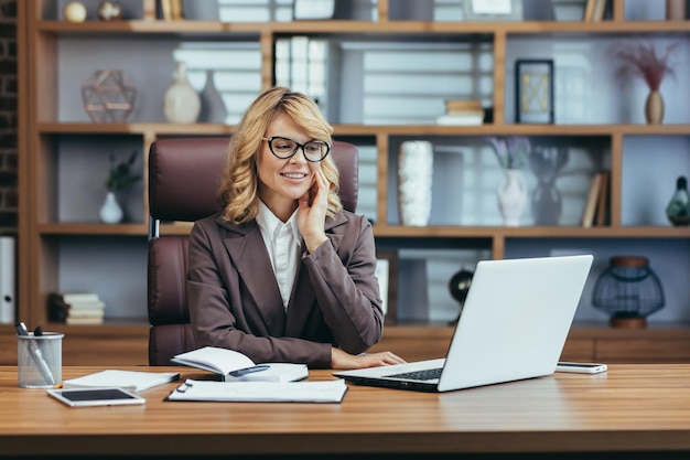 Elegante donna d'affari senior con gli occhiali che sorride mentre lavora su un computer portatile in un ambiente moderno