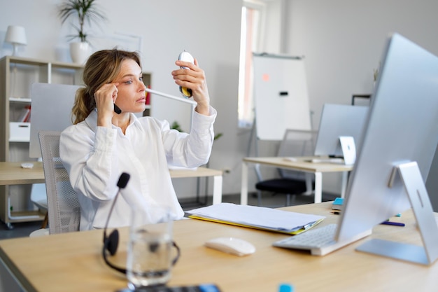 Elegante donna d'ufficio si siede al suo posto di lavoro e si guarda allo specchio per ritoccare il trucco