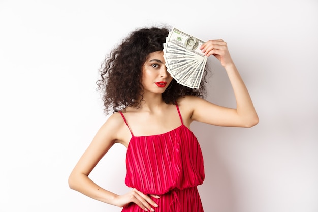 Elegante donna dai capelli ricci in abito rosso, mostrando fan del denaro e guardando fiducioso. Ragazza ricca con banconote da un dollaro, sfondo bianco.