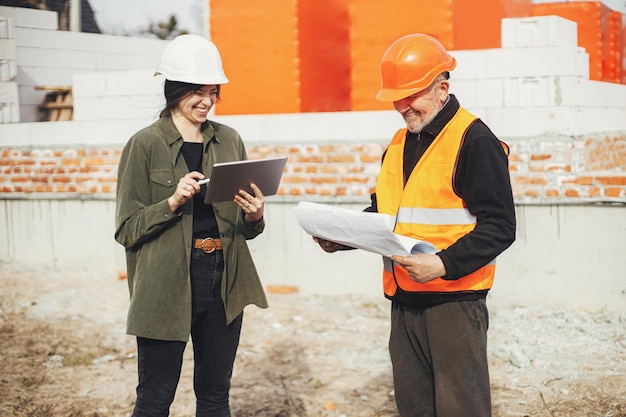 Elegante donna felice architetto con tablet e caposquadra senior che controlla i progetti in cantiere Ingegnere e operaio edile in elmetto protettivo guardando i piani della nuova casa moderna Lavoro di squadra